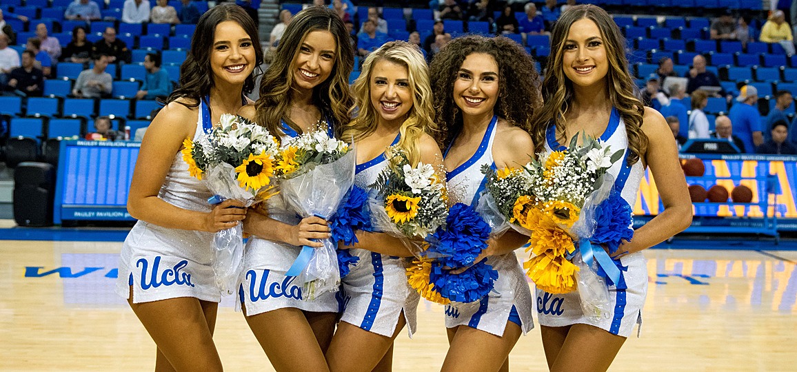Five female members of the UCLA Dance Team in white dresses, holding blue and white floral bouquets, posed in the middle of the Pauley Pavilion basketball court