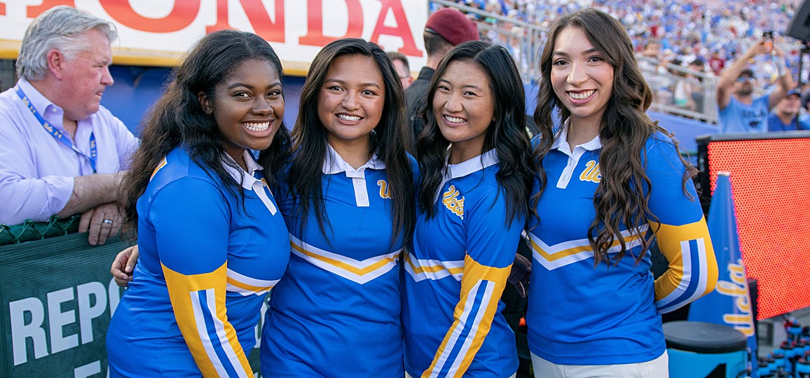 Four female members of the Yell Crew in their blue uniforms, standing in front of the gates on the field level of the Rose Bowl Stadium, fans visible in the background