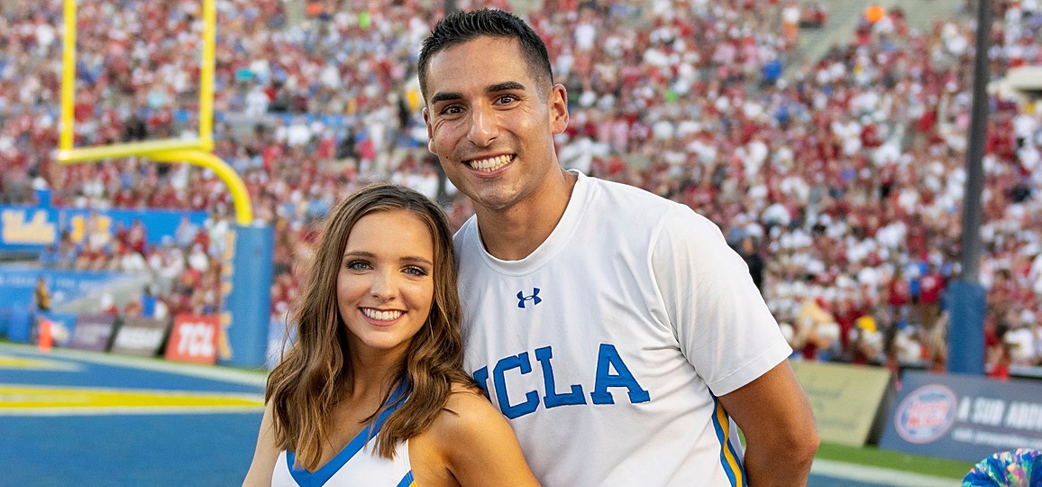 Stunting partners on the UCLA Cheer Squad (one male, one female) in white uniforms, posing on the field at Rose Bowl during the UCLA / Oklahoma football game.