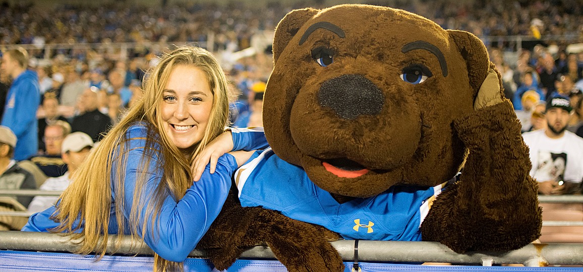 Joe Bruin sitting in the stands at the Rose Bowl with a member of the Yell Crew, both wearing blue uniforms.