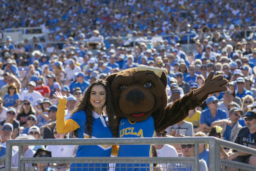 Yell Crew member in a blue uniform posed with Josie Bruin on a perch in the lower level of the Rose Bowl stadium, with thousands of UCLA fans visible behind them